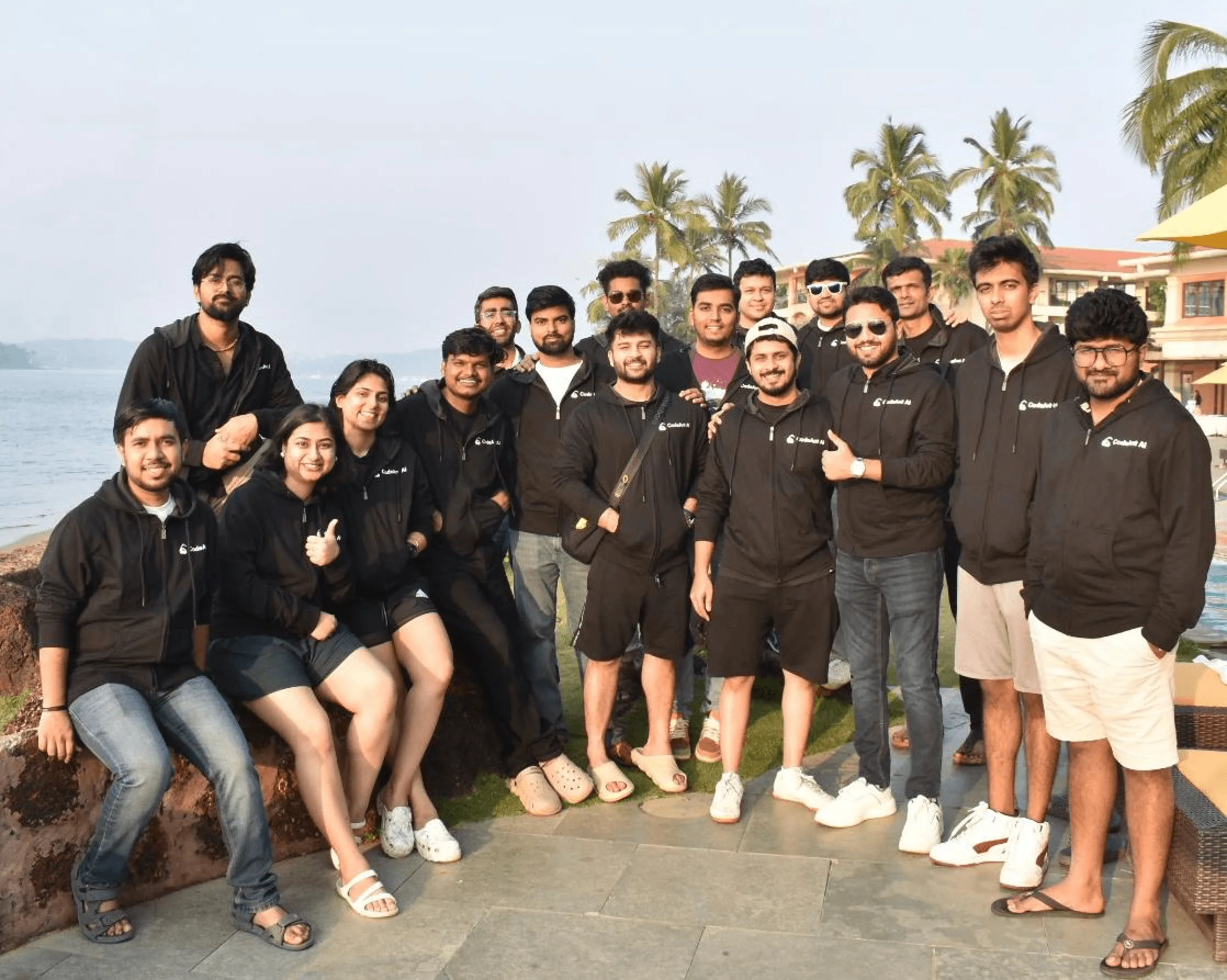 About 18 CodeAnt AI team members in matching black branded hoodies on a seaside terrace with palm trees